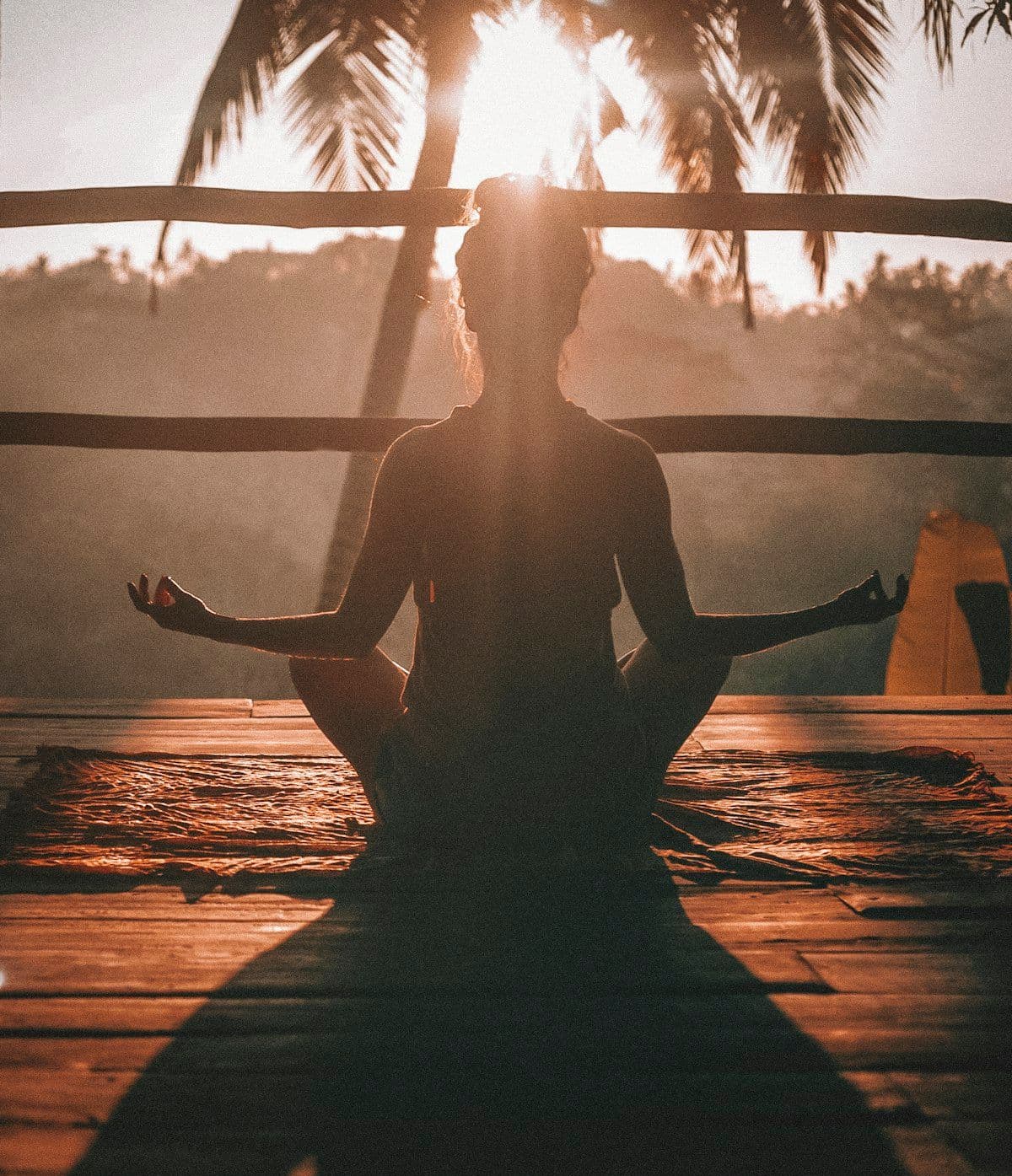 A person practising yoga at sunrise on a wooden deck framed by palms